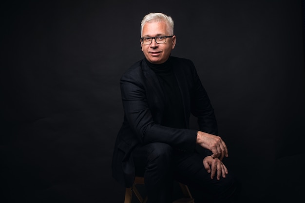 Vetri d'uso dell'uomo d'affari serio con capelli bianchi alla macchina fotografica con un'espressione messa a fuoco contro un fondo nero dello studio.