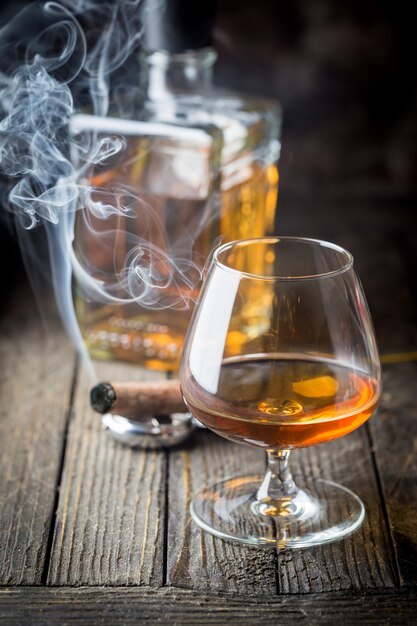 Vetro e una bottiglia di brandy o cognac e sigaro fumante sul tavolo di legno.