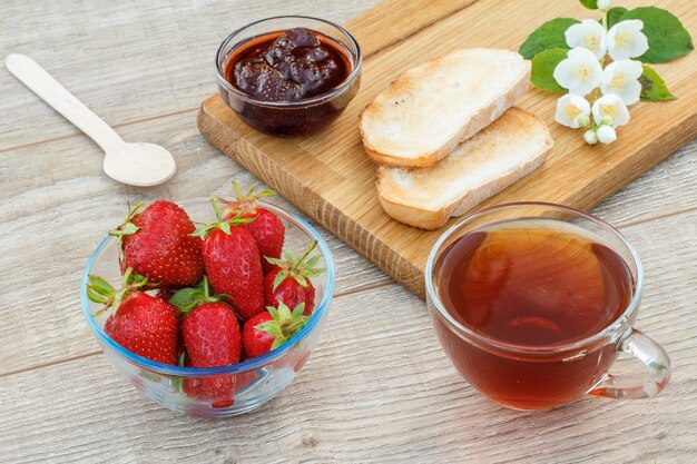 Vetro tazza di tè, marmellata di fragole fatta in casa, pane su tagliere di legno e fragole fresche, cucchiaio e fiori di gelsomino bianco su fondo di legno. Vista dall'alto.