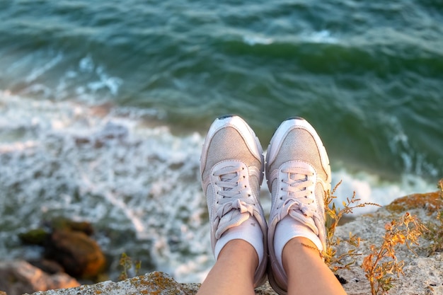 Viaggi e turismo Gambe femminili in scarpe da ginnastica bianche appese a una riva rocciosa sulla costa del mare