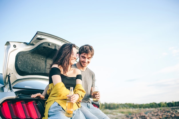 Viaggi, turismo - L'uomo e la donna bevono champagne vicino all'acqua su un tavolo portatile pieghevole. Picnic vicino all'acqua. Coppia che va in avventura. Concetto di viaggio in auto.