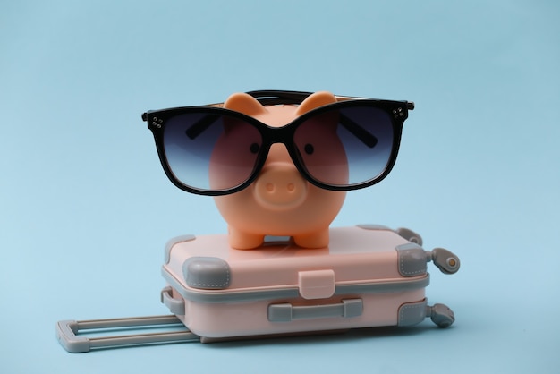 Viaggi, vacanze estive o concetto di turismo. Mini valigia da viaggio con salvadanaio in occhiali da sole su blu