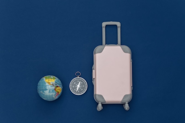 Viaggia piatto. Mini valigia da viaggio in plastica, globo, bussola su sfondo blu classico. Stile minimale. Vista dall'alto