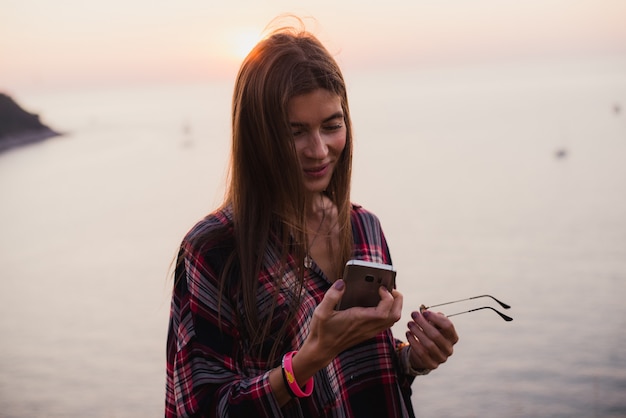 Viaggiatore alla moda della donna dei pantaloni a vita bassa sul mare al tramonto con il suo telefono cellulare