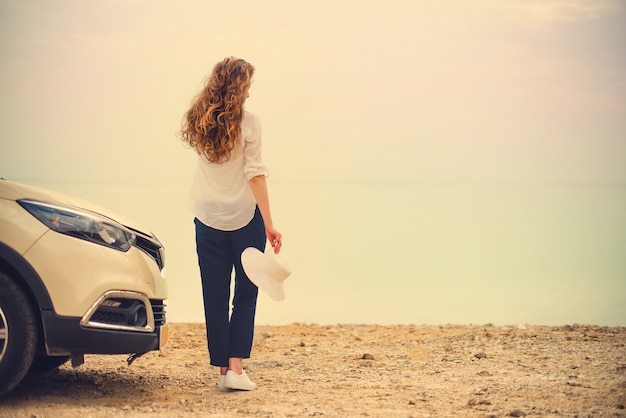 Viaggiatore alla moda felice della giovane donna sulla strada della spiaggia che si siede sull&#39;automobile bianca dell&#39;incrocio