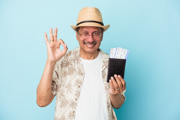 Viaggiatore anziano indiano uomo di mezza età con passaporto isolato su sfondo blu allegro e fiducioso che mostra gesto ok.