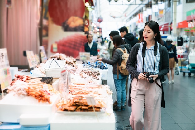 Viaggiatore asiatico della donna con la macchina fotografica che passa da una bancarella locale che vende frutti di mare trasformati giapponesi mentre visita il mercato di kuromon ichiba a Osaka in Giappone