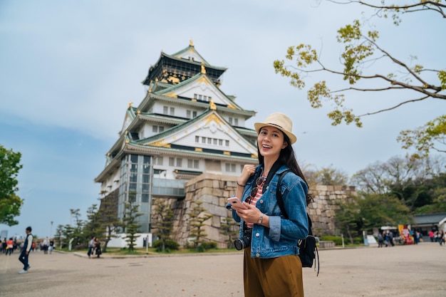 viaggiatore attraente in piedi di fronte al castello di osaka utilizzando il telefono alla ricerca del biglietto d'ingresso. giovane donna turista gioiosa fotocamera viso sorridente in giornata di sole. stile di vita da vacanza bella signora.