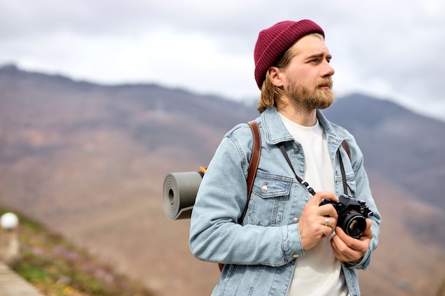 Viaggiatore che cammina in montagna con la macchina fotografica, godendosi il paesaggio, ritratto con vista laterale. Maschio caucasico barbuto in abbigliamento casual all'aperto, con zaino. Copia spazio per la pubblicità