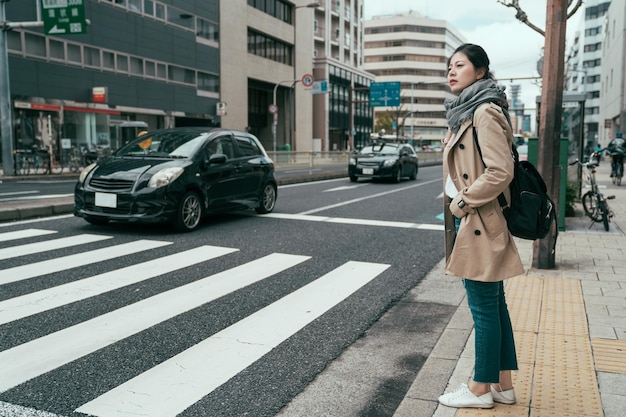 viaggiatore cinese abbastanza asiatico che cammina nella città di osaka in attesa che i semafori attraversino una strada trafficata con le auto che passano. ragazza a figura intera con borsa in piedi sulla strada e pronta a passare pedonale
