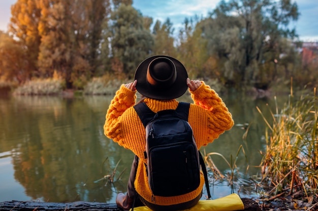 Viaggiatore con lo zaino che si rilassa dal fiume di autunno al tramonto. Giovane donna che si siede sulla banca e che si distende tenendo il cappello