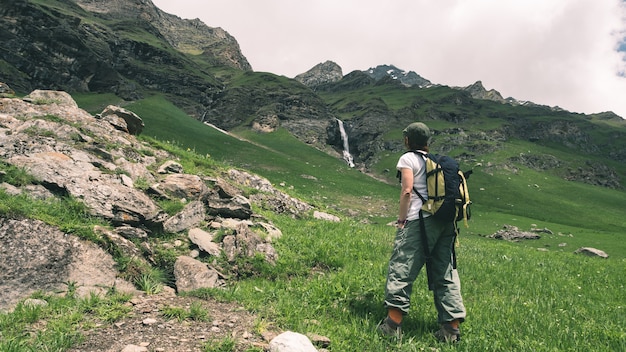 Viaggiatore con zaino e sacco a pelo della donna che fa un'escursione nel paesaggio idilliaco, nella cascata e nel prato di fioritura.