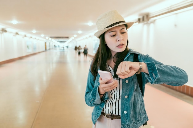 viaggiatore con zaino e sacco a pelo giovane donna asiatica in possesso di smart phone guardando il tempo sull'orologio in fretta per prendere il treno. accigliata turista femminile pendolare controlla il tempo preoccupato in ritardo. ragazza stanca che corre sottopassaggio