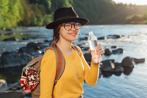 Viaggiatore della giovane donna che beve acqua pura dalla bottiglia sul fondo del fiume della natura