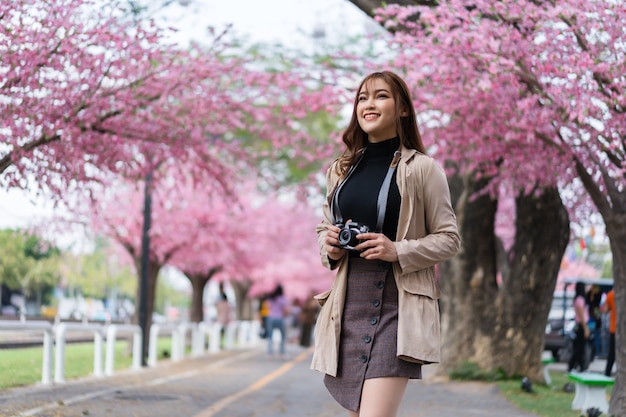 Viaggiatore della giovane donna che osserva i fiori di ciliegia o fiore di sakura che sboccia e che tiene la macchina fotografica per scattare una foto nel parco