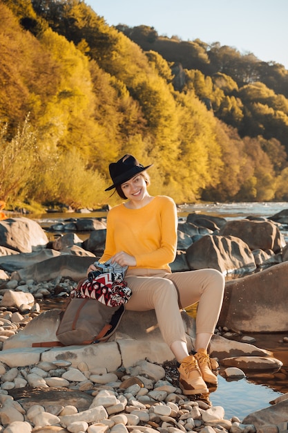 Viaggiatore della giovane donna con il telefono cellulare in mani che si siede sulla roccia vicino alla natura del fiume