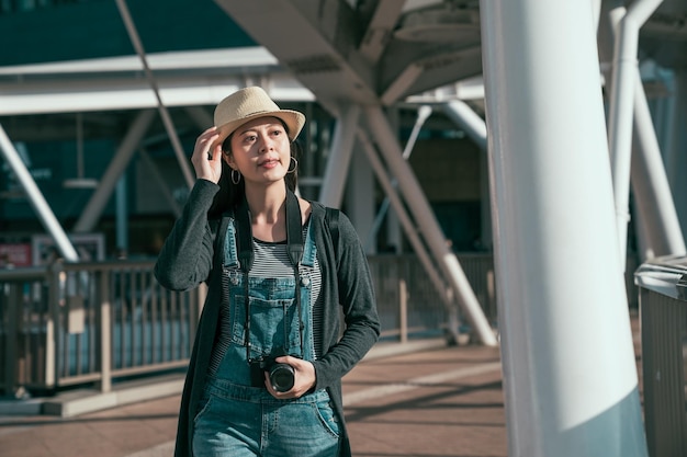 Viaggiatore di giovane donna alla moda che cammina con la macchina fotografica sulla passerella nella città di osaka. la fotografa sorridente della ragazza si gode il sole all'aperto nell'area di abeno ku. signora felice in tuta di jeans all'aperto in una giornata di sole.