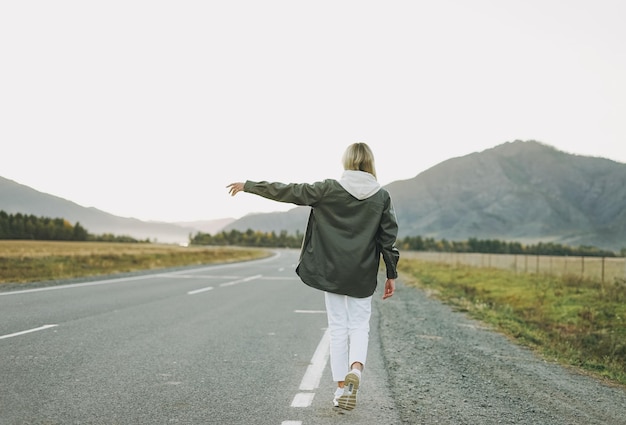 Viaggiatore di giovane donna bionda in felpa con cappuccio bianca in viaggio in montagna che fa l'autostop