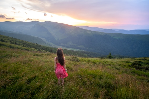 Viaggiatore di giovane donna felice in abito rosso in piedi sul pendio erboso in una serata ventosa in montagne estive godendo della vista della natura al tramonto.