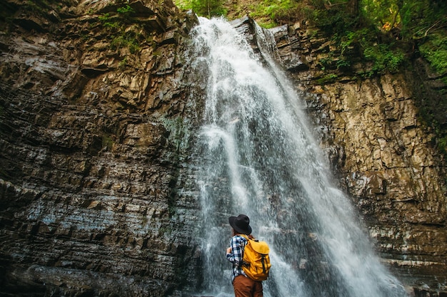 Viaggiatore donna che indossa un cappello e uno zaino giallo mentre sta in piedi contro una cascata