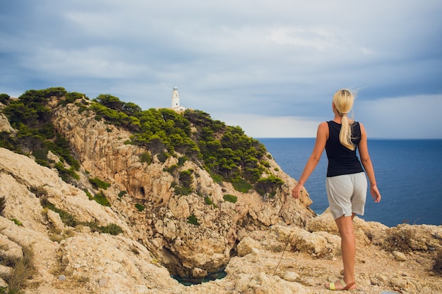 Viaggiatore giovane bella ragazza con uno zaino si trova sulla città e il faro sulla riva dell'oceano.