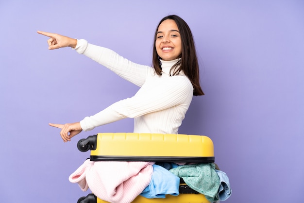 Viaggiatore giovane donna con una valigia piena di vestiti che punta il dito verso il lato e che presenta un prodotto