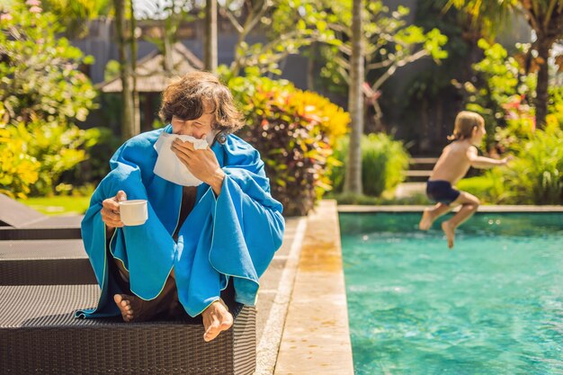 Viaggiatore malato. L'uomo ha preso un raffreddore in vacanza, siede triste in piscina a bere il tè e si soffia il naso in un tovagliolo. Suo figlio sta bene e sta nuotando in piscina. Concetto di assicurazione di viaggio.