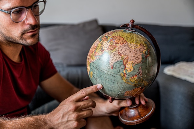 Viaggiatore maschio contentissimo che sceglie il paese sul globo del pianeta Terra prima dell'avventura mentre era seduto in camera con la valigia