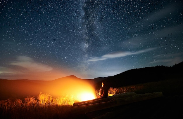 Viaggiatore maschio seduto vicino al fuoco sotto il cielo stellato notturno