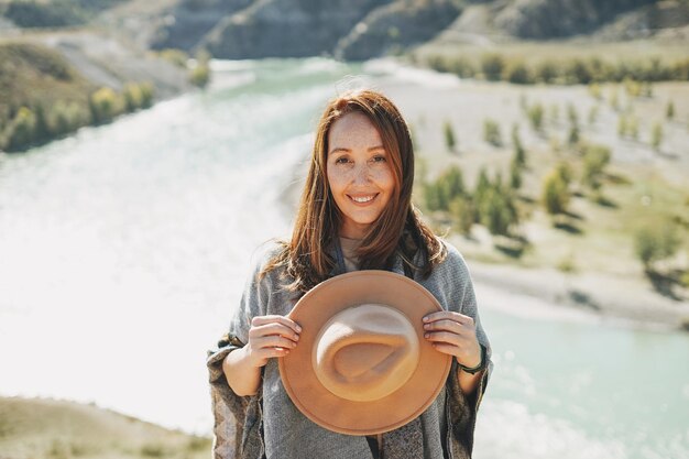 Viaggiatore spensierato della giovane donna castana in poncho con cappello di feltro sullo sfondo del fiume di montagna
