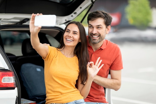 Viaggiatori di coppie felici che prendono selfie accanto al bagagliaio aperto dell'auto