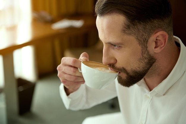 Viaggio d'affari e concetto di persone. Ritratto ravvicinato di attraente affascinante uomo d'affari caucasico di successo con la barba che beve caffè appena fatto nella camera d'albergo, per un perfetto inizio della giornata