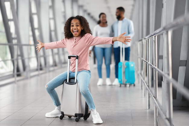 Viaggio di vacanza tanto atteso. Ragazza nera eccitata gioiosa che si siede sulla borsa e che si diverte in aeroporto. Figlia ridente che allarga le braccia e guarda in posa in telecamera, i genitori che camminano su sfondo sfocato