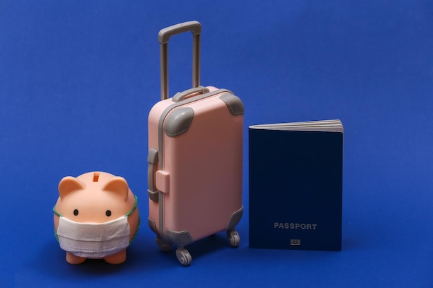 Viaggio durante il covid-19. Mini valigia da viaggio in plastica e salvadanaio con maschera facciale, passaporto su sfondo blu classico.