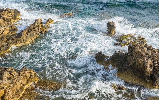 Viaggio, Estate scogliera a ridosso del Mar Mediterraneo, onde forti si infrangono con le rocce e lasciano colori blu e turchesi insieme alla schiuma del mare.