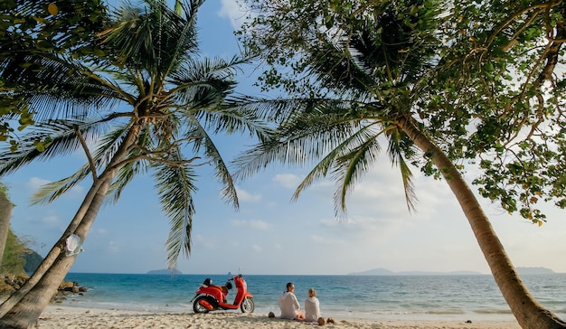 Viaggio in scooter. Bella coppia su una moto rossa in abiti bianchi sulla spiaggia di sabbia. Persone appena sposate baciano abbracci camminando vicino alle palme tropicali, al mare. Matrimonio in luna di miele sull'oceano. Noleggio moto.
