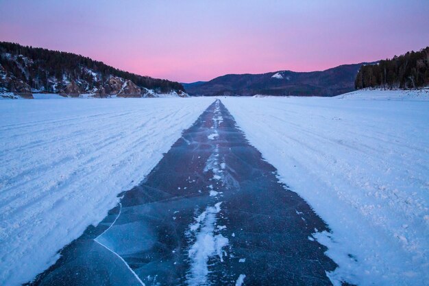 Viaggio invernale Una strada ghiacciata lungo un letto di fiume ghiacciato montagne all'orizzonte un cielo al tramonto rosa Bellezza del paesaggio invernale della natura