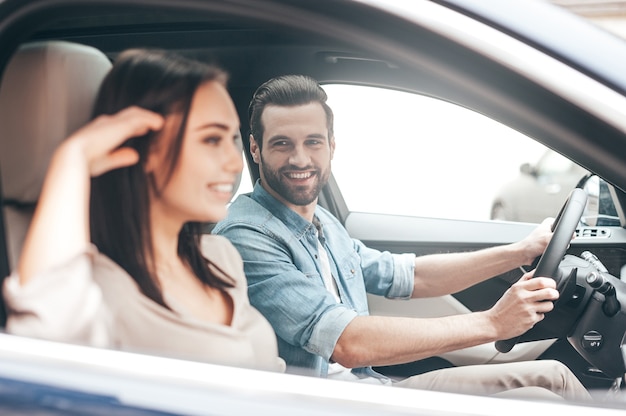 Viaggio piacevole. Bella giovane coppia seduta sui sedili del passeggero anteriore e sorridente mentre un bell'uomo si tiene per mano sul volante