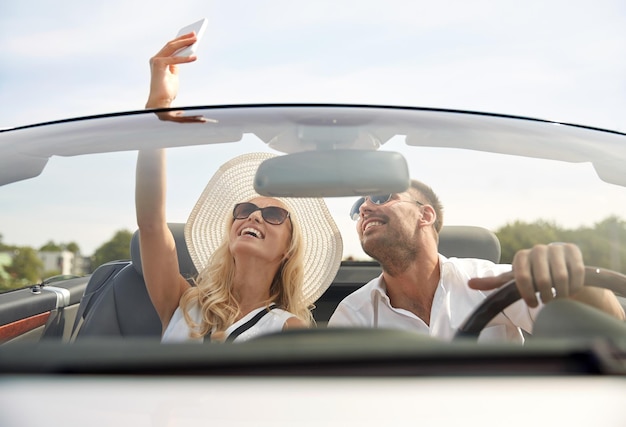 viaggio su strada, tempo libero, coppia, tecnologia e concetto di persone - uomo e donna felici che guidano in un'auto cabriolet e si fanno selfie con lo smartphone