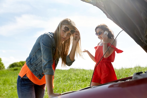 viaggio su strada, trasporto, viaggio e concetto di persone - giovani donne con cofano aperto di auto rotta in campagna che chiamano lo smartphone per il servizio di evacuazione