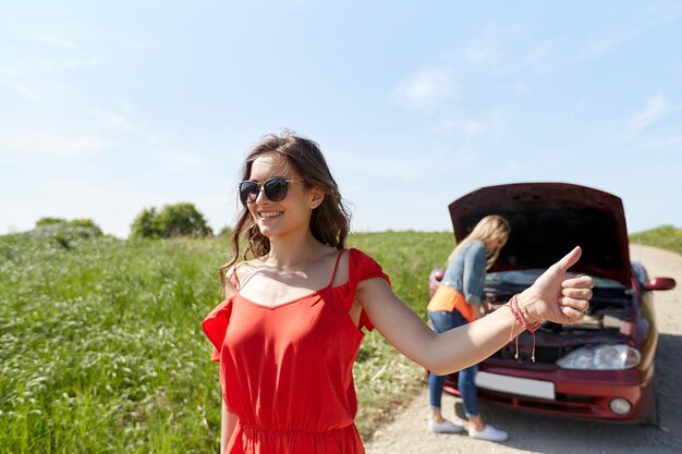 viaggio su strada, trasporto, viaggio, gesto e concetto di persone - giovani donne con auto rotta che mostrano un gesto di autostop chiedendo aiuto in campagna