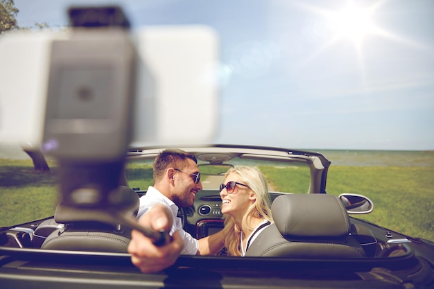 viaggio su strada, viaggio, coppia, tecnologia e concetto di persone - uomo e donna felici che guidano in auto cabriolet e scattano foto con lo smartphone sul selfie stick