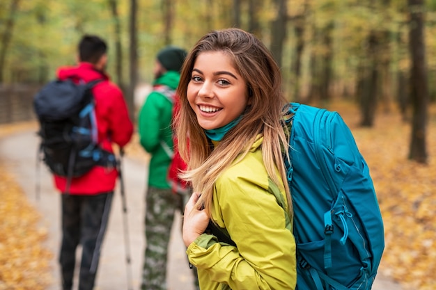 Viandante femminile allegro con lo zaino che sorride e che guarda l'obbiettivo sopra la spalla mentre cammina sul sentiero nella foresta di autunno vicino agli uomini