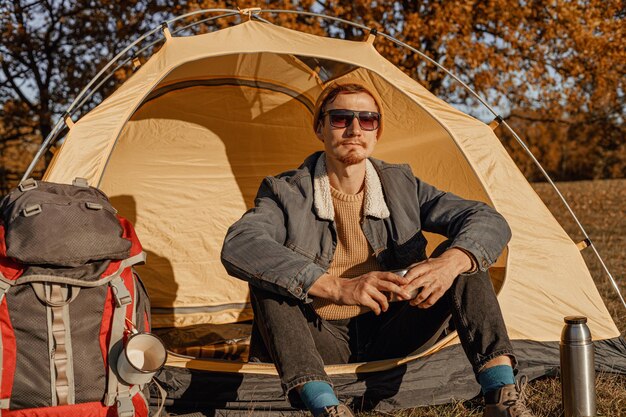 Viandante maschio che si siede vicino alla tenda al campeggio e che beve tè caldo dalla tazza di viaggio in autunno