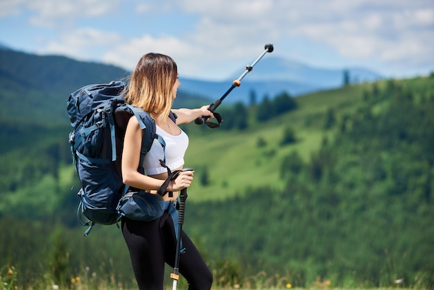 Viandante sportiva della ragazza con lo zaino che sta sulla cima di una collina, indicante via con il suo palo di trekking nelle montagne