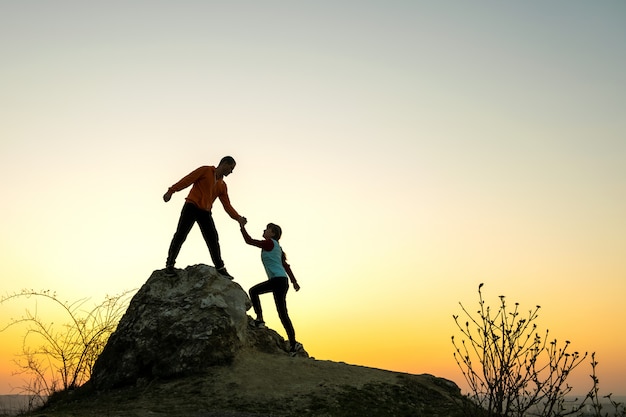 Viandanti della donna e dell'uomo che si aiutano a vicenda per scalare una grande pietra al tramonto in montagna. Coppia la scalata su un'alta roccia nella natura di sera. Concetto di turismo, viaggi e stile di vita sano.