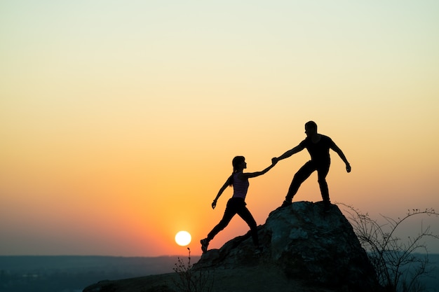 Viandanti della donna e dell'uomo che si aiutano a vicenda per scalare una grande pietra al tramonto in montagna