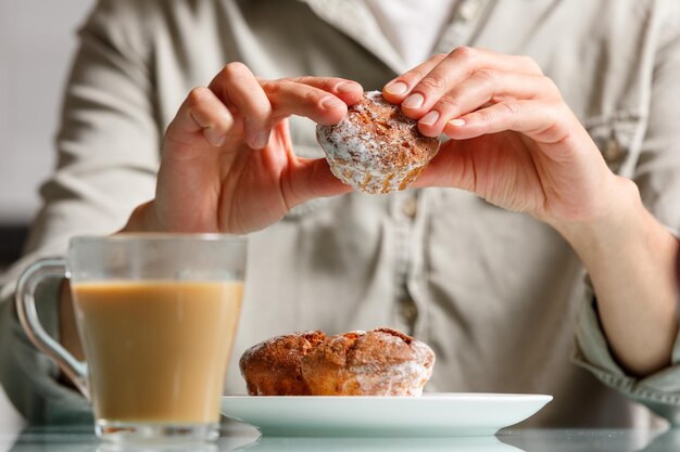 vicino di mani che tengono muffin di formaggio su un piatto con due altre torte e una tazza di caffè in vetro con latte in piedi sul tavolo