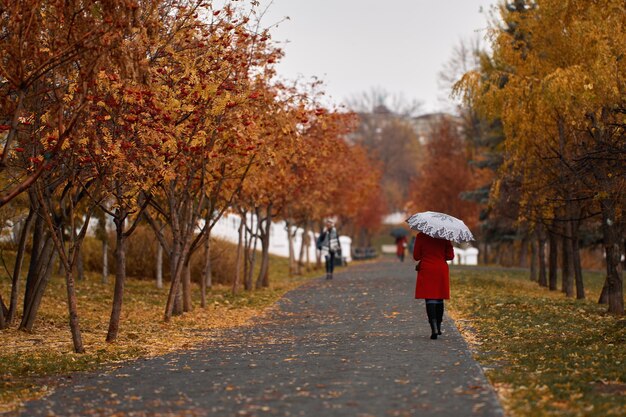 Vicolo nel parco autunnale con alberi di sorbo rosso Una donna con un cappotto rosso e un ombrello cammina lungo il vicolo autunnalexA