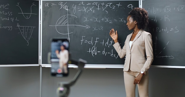 Video lezione di registrazione dello smartphone a scuola. Studio in linea. Giovane insegnante femminile afroamericana che spiega le formule di matematica o di fisica in classe. Concetto di quarantena. Studio via Internet. Isolamento.